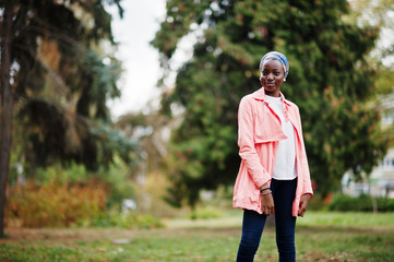 Obraz premium Young modern fashionable, attractive, tall and slim african muslim woman in hijab or turban head scarf and pink coat posed at park.
