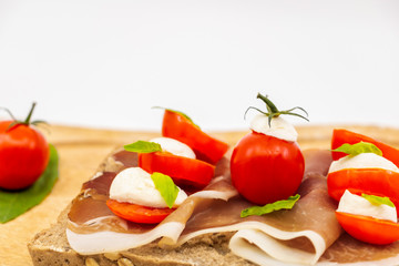 Sandwich italien sur une tranche de pain multi céréales avec une tranche de jambon rouge des tomates cerises billes de mozzarella et du basilic frais sur une assiette en bois -isolé fond blanc