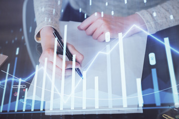 Hand taking notes in notepad. Forex chart holograms in front. Concept of research. Multi exposure