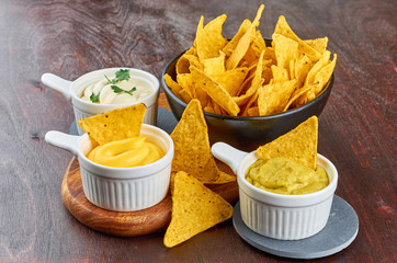 Nachos - yellow corn chips with various sauces in bowls: guacamole, cheese sauce, white sauce, on a wooden table. Mexican food concept.