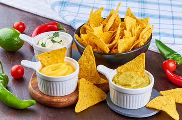 Nachos - yellow corn chips with various sauces in bowls: guacamole, cheese sauce, white sauce, on a wooden table. Mexican food concept.