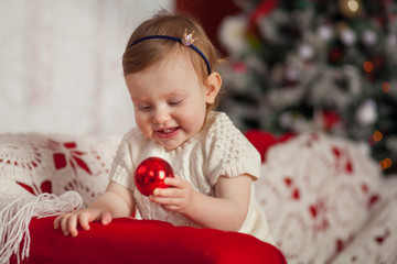 Fototapeta na wymiar Portrait of a cute little girl dressed in a knitted white dress holding a red Christmas ball in a Christmas interior with lights. Christmas and new year concept.