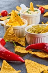 Nachos - yellow corn chips with various sauces in bowls: guacamole, cheese sauce, white sauce, on dark background. Mexican food concept.