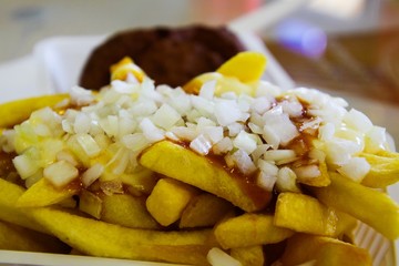 Frikandel with pommes spezial: View on heap French fries covered with fat sticky mayonnaise, ketchup and onions in white plastic bowl. Blurred meat ball background. Netherlands