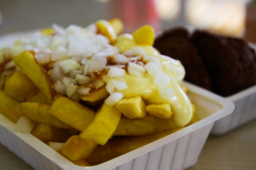 Frikandel with pommes spezial: View on heap French fries covered with fat sticky mayonnaise, ketchup and onions in white plastic bowl. Blurred meat ball background. Netherlands