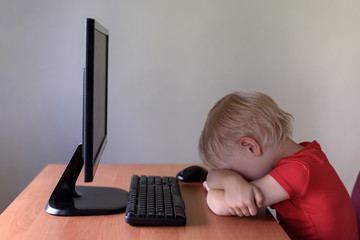 Tired little blond boy sleeping on a table under the monitor PC. Internet and preschooler