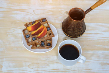 Chocolate Belgian waffles with fruits, cup of coffee and cezve on white wooden background. Delicious breakfast. Top view