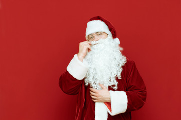 Portrait of a beautiful Santa Claus, standing on a red background and posing at the camera, wearing a classic red suit and beard, twirling his mustache. Man in santa costume isolated on red background