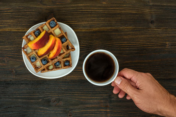 Belgian waffles with fruit and berries. Male hand with cup of coffee. Top view