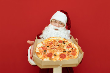 Smiling Santa Claus stands on a red background with a box of mouth-watering hot pizza in her hands and points it into the camera. Santa delivers pizza for Christmas, looks into the camera with a smile