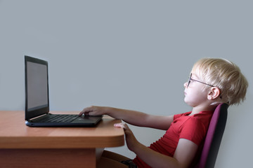 Funny blond boy in glasses sits at a laptop. Internet and preschooler