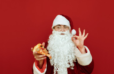 Fototapeta na wymiar Handsome Santa with a slice of pizza in her hand stands on a red background, looks in camera and shows her thumbs sign OK. Man in a Santa suit eats a pizza. Pizza for Christmas