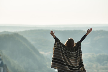 Young woman enjoying sunny day on a hill. Raised hands. Back view