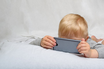Little blond boy looking something his nose buried in a smartphone, lying in bed. Gadget Leisure