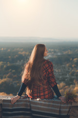 Young woman with long hair sits on a hill overlooking the village. Back view