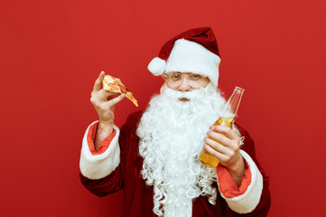 Happy Santa with a bottle of beer and a piece of pizza in his hands stands on a red background, looks into the camera and smiles. Santa drinks alcohol and eats fast food. Christmas.