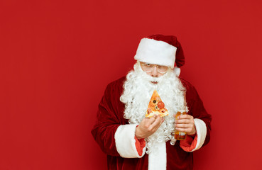 Portrait of an evil Santa Claus drinking alcohol on a red background with a beer bottle and a piece of pizza in his hand,with an evil face looking into the camera. Bad Santa isolated on red background