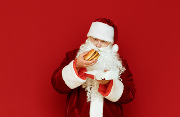 Fototapeta na wymiar Funny man in Santa Claus costume stands on red background with burger in hand, looks at fast food with surprised face. Shocked Santa isolated on red background with sandwich in hand.