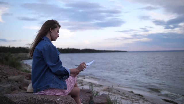 A young woman artist makes a sketch in pencil on an album. The girl is sitting on the seashore. 4K Slow Mo