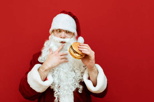 Funny man in Santa Claus costume stands with a burger in his hands on a red background,thoughtfully looking at fast food.Cheerful Santa eating fast food. Christmas and New Year. Harmful food