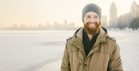 Close-up Caucasian young male red hair and beard in a hat and a park coat posing winter model against a background of a lake to freeze snow © Elizaveta
