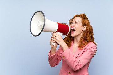 Redhead woman in suit over isolated blue wall shouting through a megaphone