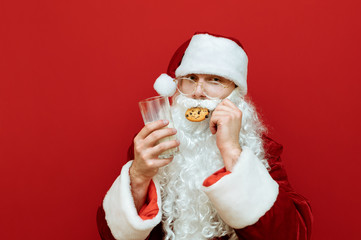 Man in a Santa Claus costume stands on a red background with a glass of milk in his hand and eats a chocolate chip cookie, looking in camera with a serious face. Santa loves Christmas cookies and milk
