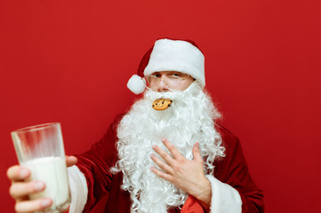 Close up portrait of a funny Santa Claus eating a delicious Christmas cookie and holding glass of milk, looking in camera and scratching his beard.Funny Santa isolated on red background. New Year