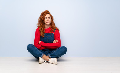Redhead woman with overalls sitting on the floor with confuse face expression