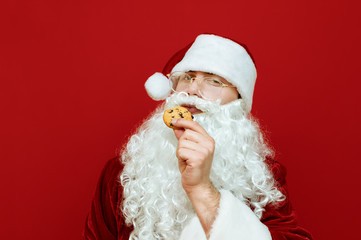 Portrait of beautiful Santa Claus with beard and glasses holding Christmas cookies in his hand and looking into the camera with a serious face. Tradition with Santa and cookies. Christmas concept.