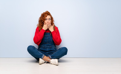 Redhead woman with overalls sitting on the floor covering mouth and looking to the side