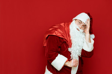 Tired Santa Claus with a bag on his back stands on a red background and looks into the camera, Santa is tired of carrying presents. Delivery of Christmas gifts by Santa Claus. Xmas concept