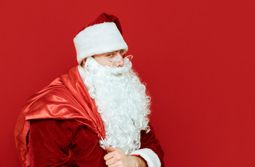 Sneering Santa Claus with bag of gifts stands on red background and looks into camera, closeup portrait. Isolated. Cunning Santa for Christmas. X mas concept. Copyspace