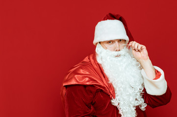 Closeup portrait of Santa Claus on red background holding a bag with gifts and looking into the camera with a serious face. Empty space to advertise on the left. Christmas and New Year concept