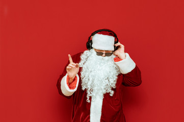Studio portrait of Santa Claus in sunglasses listening to music in headphones and dancing on a red background. Santa party-goer in headphones isolated on red background. Copyspace. Xmas concept