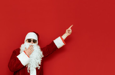 Fototapeta na wymiar Shocked Santa points her hand to a blank spot and with a surprised face looks into the camera, covered her mouth with her hand, wears sunglasses. Santa is pointing her finger at Copyspace. Christmas