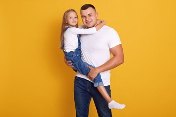Studio shot of charming kid girl with her father, handsome man holding child in hands, wearing casual clothing, posing isolated over yellow background Family, lifestyle and happyness concept.