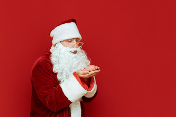Fototapeta na wymiar Portrait of Santa standing on red background looks at raised hands and sends an air kiss, looks away. Christmas concept. Happy xmas. Copyspace