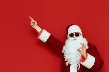 Fototapeta na wymiar Cheerful Santa Claus stands on a red background and points his hand to a blank space and with his fingers gesture ok, looks into the camera and smiles. Positive Santa pointing finger at Copyspace