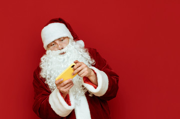 Portrait of Santa Claus gamer playing video games on smartphone on red background, looking carefully at smartphone screen. Christmas New Year gaming. Santa is playing mobile games. Copyspace