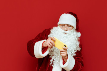 Portrait of Santa Claus gamer playing mobile games on red background, looking intently at smartphone screen. Mobile gaming and man in Santa Claus costume. X mas concept
