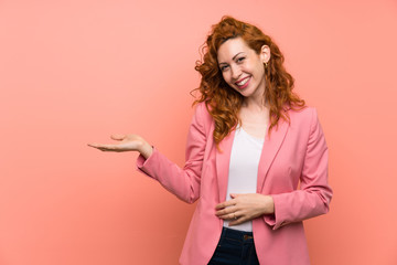 Redhead woman in suit over isolated pink wall holding copyspace imaginary on the palm