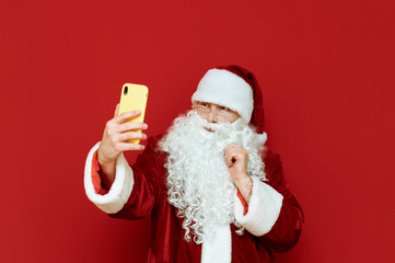 Portrait of Santa Claus taking selfie on red background, serious face looking into camera. Funny Santa with smartphone in hand isolated on red background. Copyspace. X mas concept