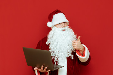 Santa Claus stands on a red background with a laptop in his hand, shows his thumb up and looks into the camera with a serious face. Business Santa working on laptop isolated. Copyspace