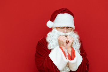 Portrait of upset Santa Claus on red background, warming from cold. Santa froze and looks into the camera with a sad face. Isolated.