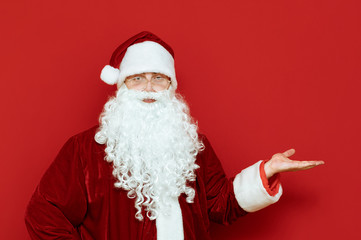 Fototapeta na wymiar Portrait of positive santa claus isolated on red background shows his hand on a blank space and smiles. Funny Santa stands on red background and shows her hand on copyspace. Xmas concept