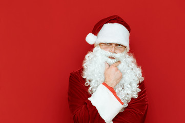 Studio portrait of a pensive santa on a red background, looks into the camera with a serious face and scratches his beard. Serious Santa thinks close portrait. Isolated. Copyspace