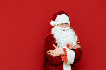 Santa Claus freezes, warms up against a red background and looks in camera with a sad face.Portrait Santa Claus who is cold. Man in santa costume freezing on red background. Cold winter for Christmas