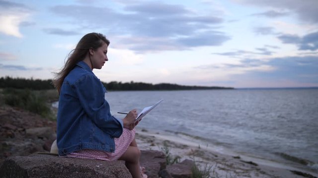 A young artist is looking for inspiration on the seashore. A girl paints a landscape in an album.4K Slow Mo