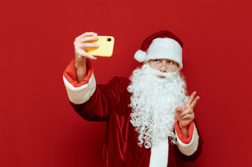 Studio photo of Santa Claus posing on smartphone camera, taking selfies and showing peace gesture to camera on red background. Selfies. Santa and technology. Merry Christmas.